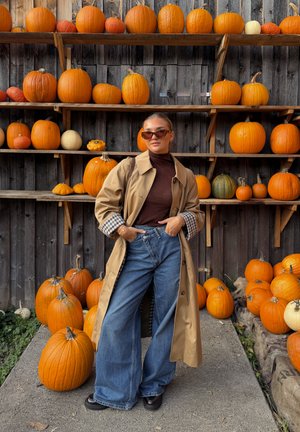 Beige trenchcoat met een geruite mouw, gecombineerd met donkere jeans. Omgeven door pompoenen in verschillende tinten, tegen een houten achtergrond.