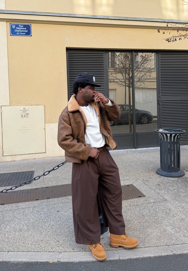 Man in brown shearling jacket, wide trousers, and tan boots stands on street near black bin and building with shuttered windows.