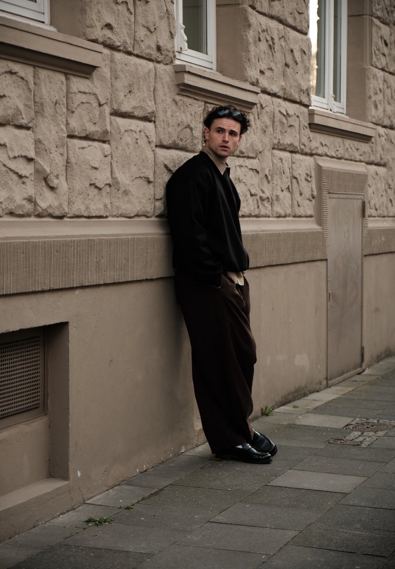 Jeune homme portant une veste noire et un pantalon foncé appuyé contre un mur en pierre texturé sur un trottoir de la ville, regardant sur le côté.