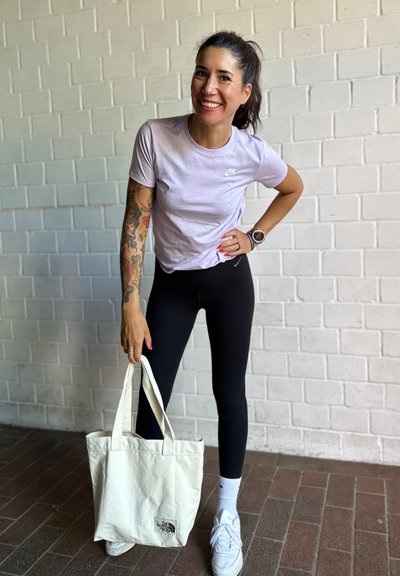 Camiseta Nike de color morado claro, leggings negros, zapatillas blancas y un bolso de mano crema con un logo. Suelo de ladrillos, fondo de pared de ladrillos blancos.