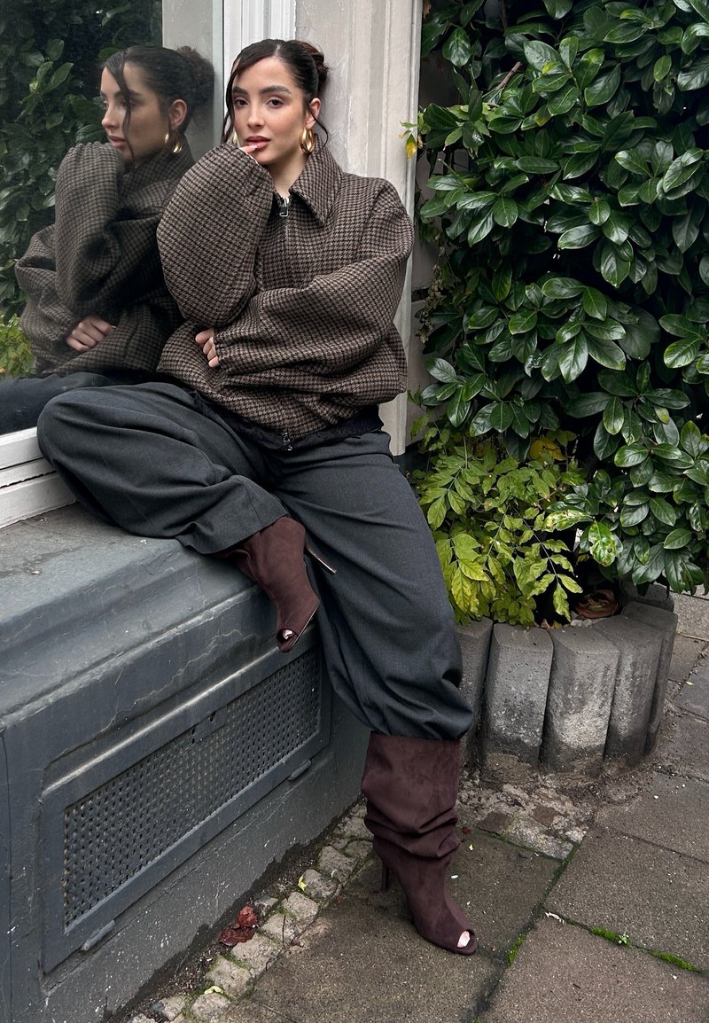 Brun rutete oversized jakke, grå bukser med vide ben og vinrøde, slappe hælede støvler. Grønn løvverk i bakgrunnen. Reflekterende vindusdetalj.