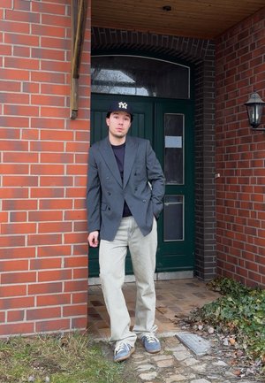 Joven con blazer informal, vaqueros claros, zapatillas deportivas y gorra de béisbol apoyado contra una pared de ladrillos junto a una puerta verde y una lámpara exterior.