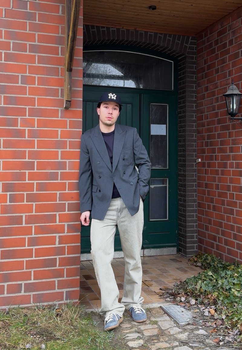 Jeune homme en blazer décontracté, jean clair, baskets et casquette de baseball, appuyé contre un mur en brique près d'une porte verte et d'une lampe extérieure.