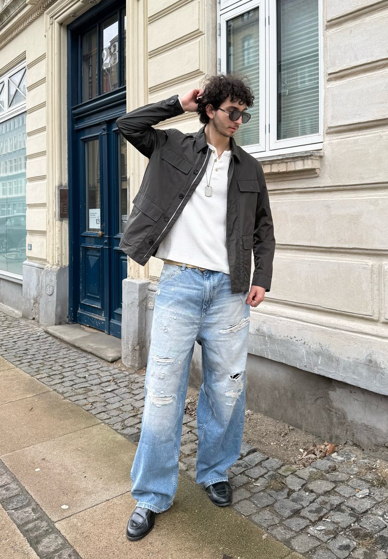 Jeune homme portant des lunettes de soleil, une veste sombre, une chemise blanche et un jean large déchiré, debout sur un trottoir en pavés près d'un bâtiment beige avec une porte bleue.