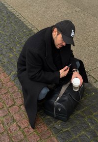 Haină neagră din lână, așezată peste un bărbat care stă ghemuit pe pavele. Ține în mână un smartphone și o ceașcă de cafea, lângă o geantă mare din piele neagră.