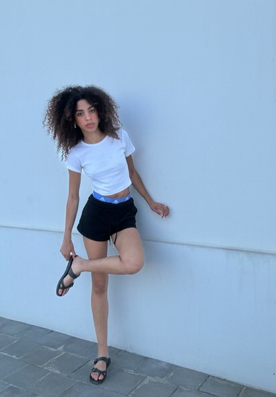 Camiseta blanca de corte cropped, pantalones cortos negros con cordón y sandalias negras. El modelo posa frente a una pared gris claro, mostrando un atuendo casual de verano.