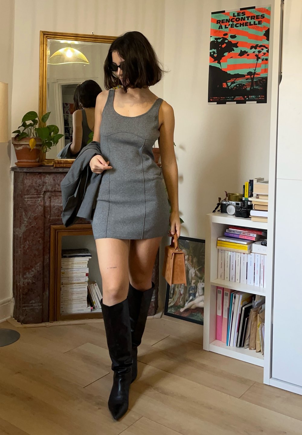 Szara sukienka bez rękawów o dopasowanym kroju z przeszyciami, zestawiona z wysokimi czarnymi kozakami i brązową torebką.