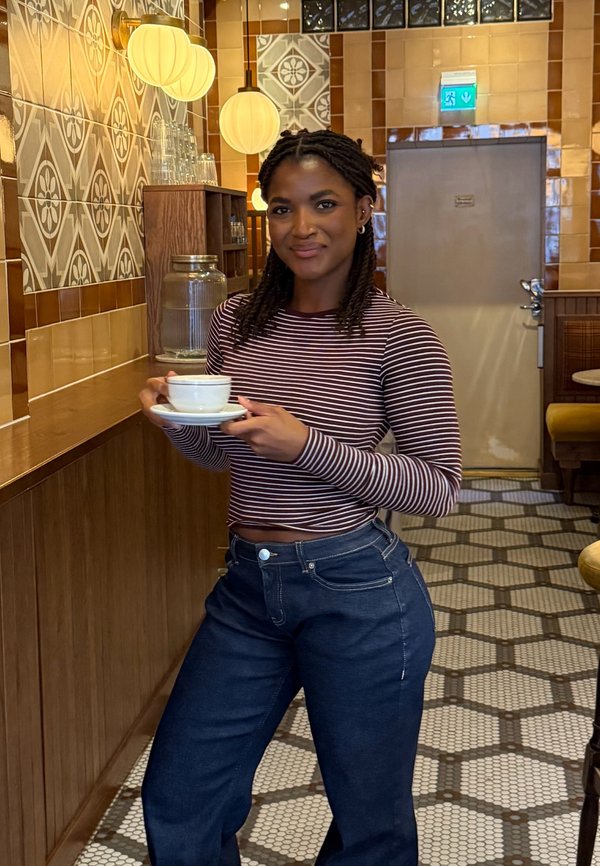 Letitettyä tukkaa oleva nainen pitelee kuppia ja lautasliinaa kuviollisella laattakahvilassa, jossa on lämmin valaistus ja puinen sisustus.