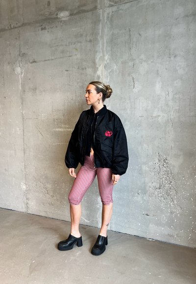 Chaqueta bomber negra con un bolsillo frontal, detalle de cereza bordado en rojo, combinada con leggings de cuadros rojos y blancos y zapatos de plataforma negros y gruesos.