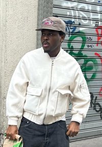 Bílá bomber bunda se zipem a několika kapsami, šedá čepice s logem, černé džíny, stojící proti zdi pokryté graffiti.