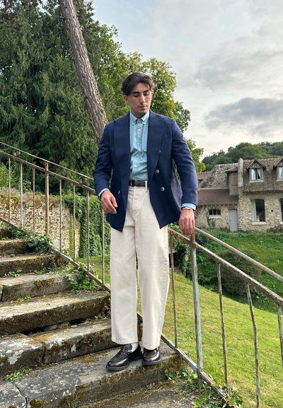 Blazer azul marino, camisa de botones azul claro, pantalones color crema; zapatos negros. Entorno al aire libre con escalones de piedra y vegetación de fondo.