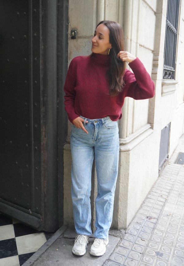 Une femme en pull bordeaux, jean bleu clair et baskets blanches se tient près d'une porte, tenant ses cheveux.