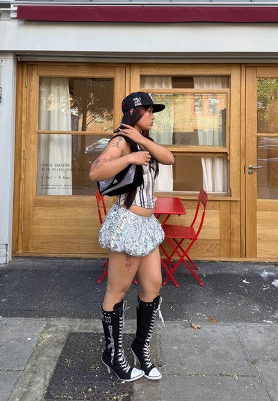Gorra negra con texto, top sin mangas a rayas, mini falda plisada plateada brillante y botas negras de cuero altas hasta la rodilla con punteras blancas.