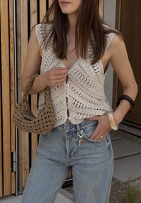 Háčkovaná béžová top bez rukávů s výstřihem do V, zdobený složitými vzory a knoflíky. V kombinaci se světle modrými džíny s vysokým pasem a tkanou hnědou taškou.