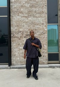 Camicia a maniche corte a righe, pantaloni scuri larghi e scarpe nere. Porta una borsa a tracolla nera e sta appoggiato a un muro grigio con texture.