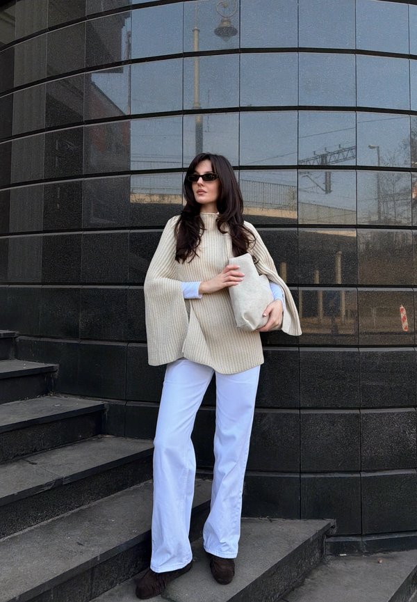 Donna con poncho beige lavorato a maglia, pantaloni bianchi, occhiali da sole neri, tiene una borsa beige, in piedi su scalini esterni davanti a una parete piastrellata nera.