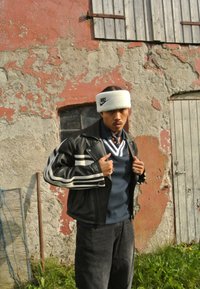 Juodas odinis švarkas su baltais dryžiais, tamsiai mėlynas marškinėliai ir balta pūkuota Nike galvos juosta. Modelis pozuoja prieš nusidėvėjusią sieną ir duris.