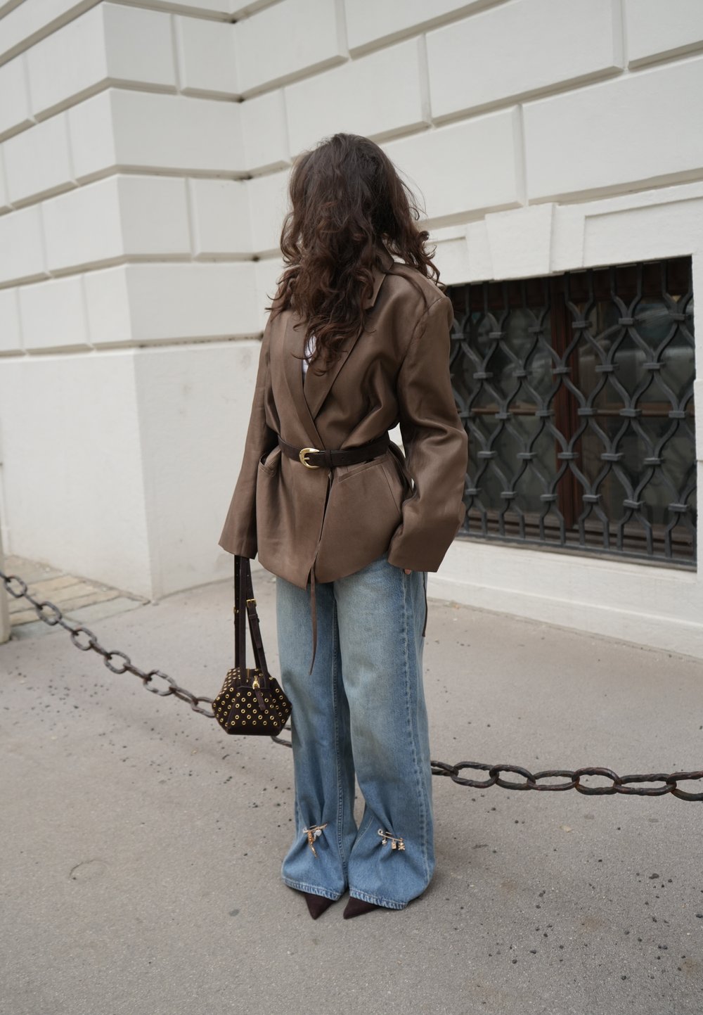 Blazer oversize en cuir marron avec une ceinture à la taille, associé à un jean bleu à jambes larges avec des accents, et un petit sac à main imprimé.