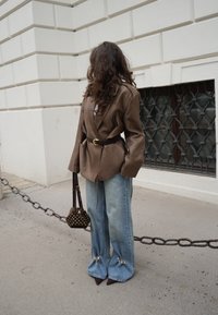 Blazer oversize en cuir marron avec une ceinture à la taille, associé à un jean bleu à jambes larges avec des accents, et un petit sac à main imprimé.