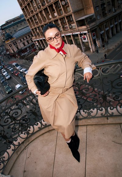 Mujer con gabardina beige y bufanda roja apoyada en una barandilla ornamentada de balcón que mira a una calle urbana concurrida con vehículos y edificios históricos.