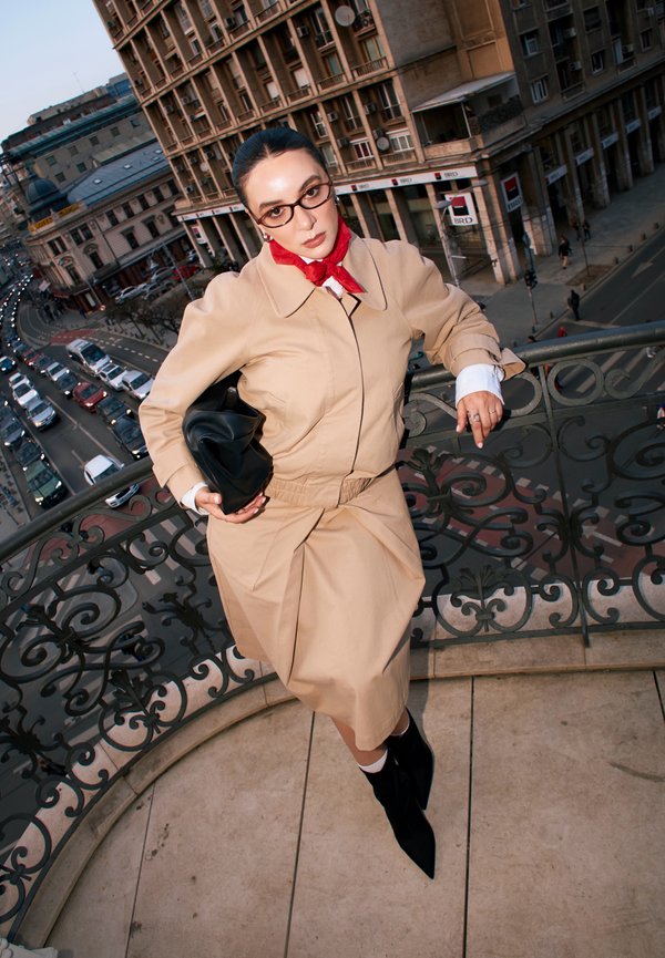 Frau in beige Trenchcoat und rotem Schal lehnt an einem verzierten Balkonengeländer und blickt auf eine belebte städtische Straße mit Fahrzeugen und historischen Gebäuden.