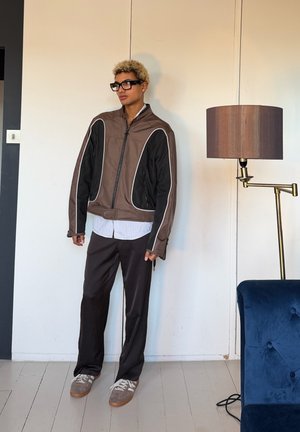 Persona con cabello corto y rizado rubio y gafas negras lleva una chaqueta marrón y negra, pantalones oscuros, zapatillas grises, de pie junto a una lámpara de pie y una silla azul.