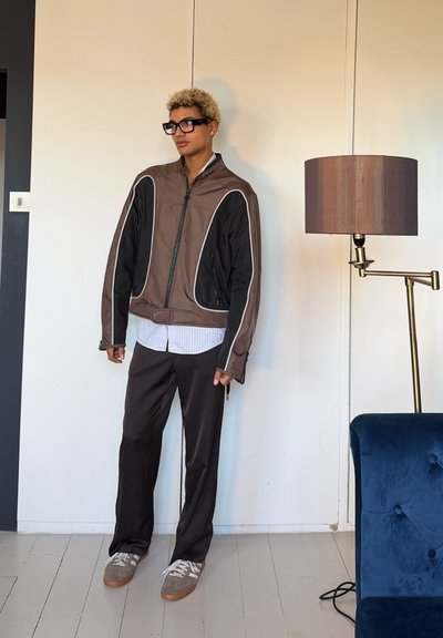 Persona con cabello corto y rizado rubio y gafas negras lleva una chaqueta marrón y negra, pantalones oscuros, zapatillas grises, de pie junto a una lámpara de pie y una silla azul.