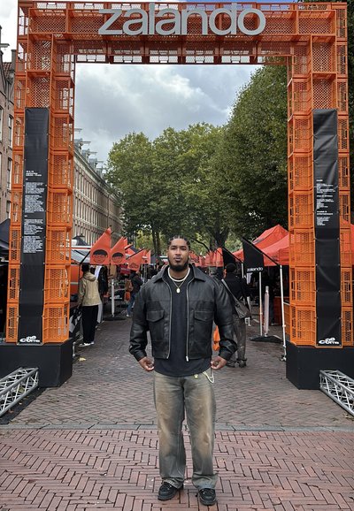 Chaqueta de cuero negra, suéter negro y pantalones gris claro. Arco naranja con cartel de "zalando". Evento al aire libre con puestos de mercado y carpas.