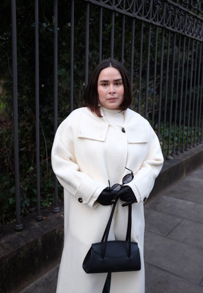 Mujer con abrigo blanco y guantes negros que sostiene un bolso negro y gafas de sol, parada junto a una cerca metálica en una acera.