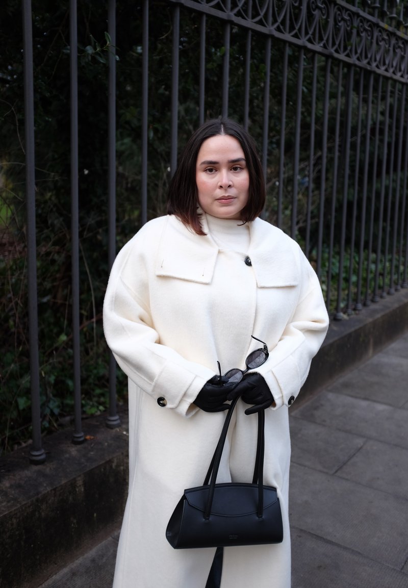 Femme en manteau blanc et gants noirs tenant un sac à main noir et des lunettes de soleil, debout près d'une clôture métallique sur un trottoir.