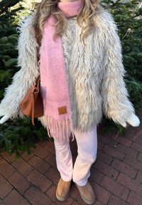 Fluffy cream faux fur jacket, light pink scarf with fringes, tan leather bag, pink trousers, and beige slip-on boots against greenery.