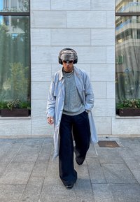 Ανοιχτό γκρι μακρύ παλτό πάνω από ένα γκρι πουλόβερ, μαύρο παντελόνι σε φαρδιά γραμμή, καπέλο με γκρι σχέδιο και ακουστικά. Τα αξεσουάρ περιλαμβάνουν κολιέ και δαχτυλίδια.