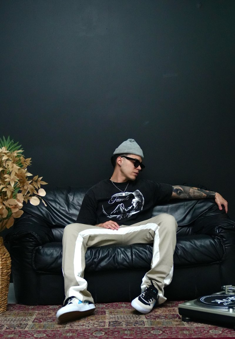 Schwarzes Ledersofa, hellbeige Hose mit weißen Streifen, schwarzes Grafik-T-Shirt, graue Mütze, Sonnenbrille, tätowierter kurzer Arm, Plattenspieler.