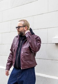 Burgundska usnjena jakna s prsnih žepih, z zadrgo in gladko teksturo, prekrita s črnim hoodijem in modrimi jeans hlačami.