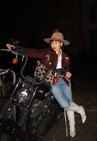 Donna con cappello beige, giacca marrone, stivali bianchi e borsa a stampa mucca seduta su una moto nera di notte in una strada cittadina.