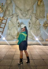 Una persona in un abbigliamento verde e nero con lunghe trecce, che indossa occhiali da sole e un cappellino, si trova di fronte a un murale che raffigura figure robotiche.
