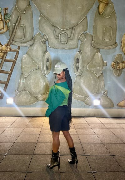 Una persona vestida con un traje verde y negro, con trenzas largas, usando gafas de sol y una gorra, está de pie frente a un mural que presenta figuras robóticas.