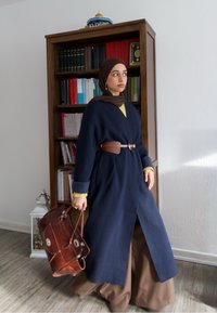 Long manteau bleu marine avec ceinture, superposé à un haut jaune et un pantalon marron. Elle porte un sac en cuir marron. En arrière-plan, des étagères remplies de livres.