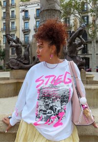 Sudadera blanca con texto gráfico en rosa y una impresión fotográfica en blanco y negro. La modelo lleva una falda de capas y un bolso rosa con un charm.