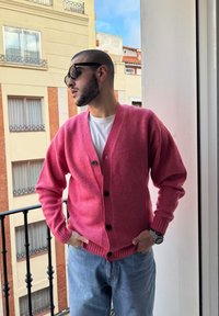 Rózsaszín gyapjú kardigán V nyakkivágással, fekete gombokkal és bordás mandzsettával, világos kék farmernadrággal párosítva, egy erkély háttérrel.
