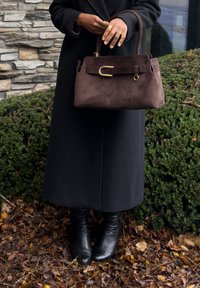 Borsa in camoscio marrone con tracolla in pelle marrone, caratterizzata da un dettaglio in metallo dorato. Sorretto da una persona con un lungo cappotto nero e stivali.