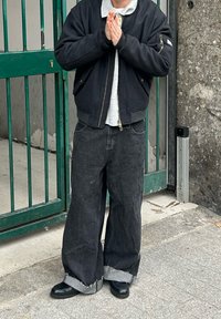 Giacca bomber nera sopra una maglietta grigia, abbinata a jeans neri a gamba larga con orli sfrangiati e stivali neri. Cancello verde sullo sfondo.