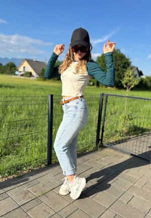 Camisa de manga larga en tonos crema y verde, jeans azul claro remangados y deportivas blancas gruesas, estilizadas con una gorra negra y gafas de sol.
