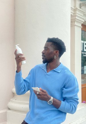 Botella de bomba blanca sostenida en una mano; el envase en la otra. La persona lleva puesto un suéter de canalé azul y joyería de plata, con un reloj en la muñeca.