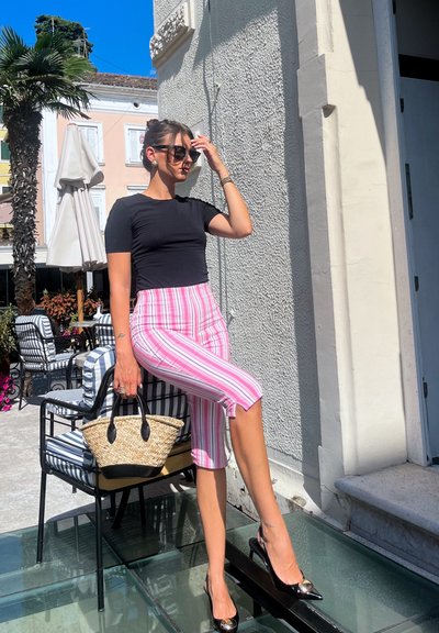 Top negro ajustado, pantalones capri a rayas rosas, tacones negros de punta y un bolso de fibra trenzada. De pie al aire libre con palmeras y sillas de fondo.