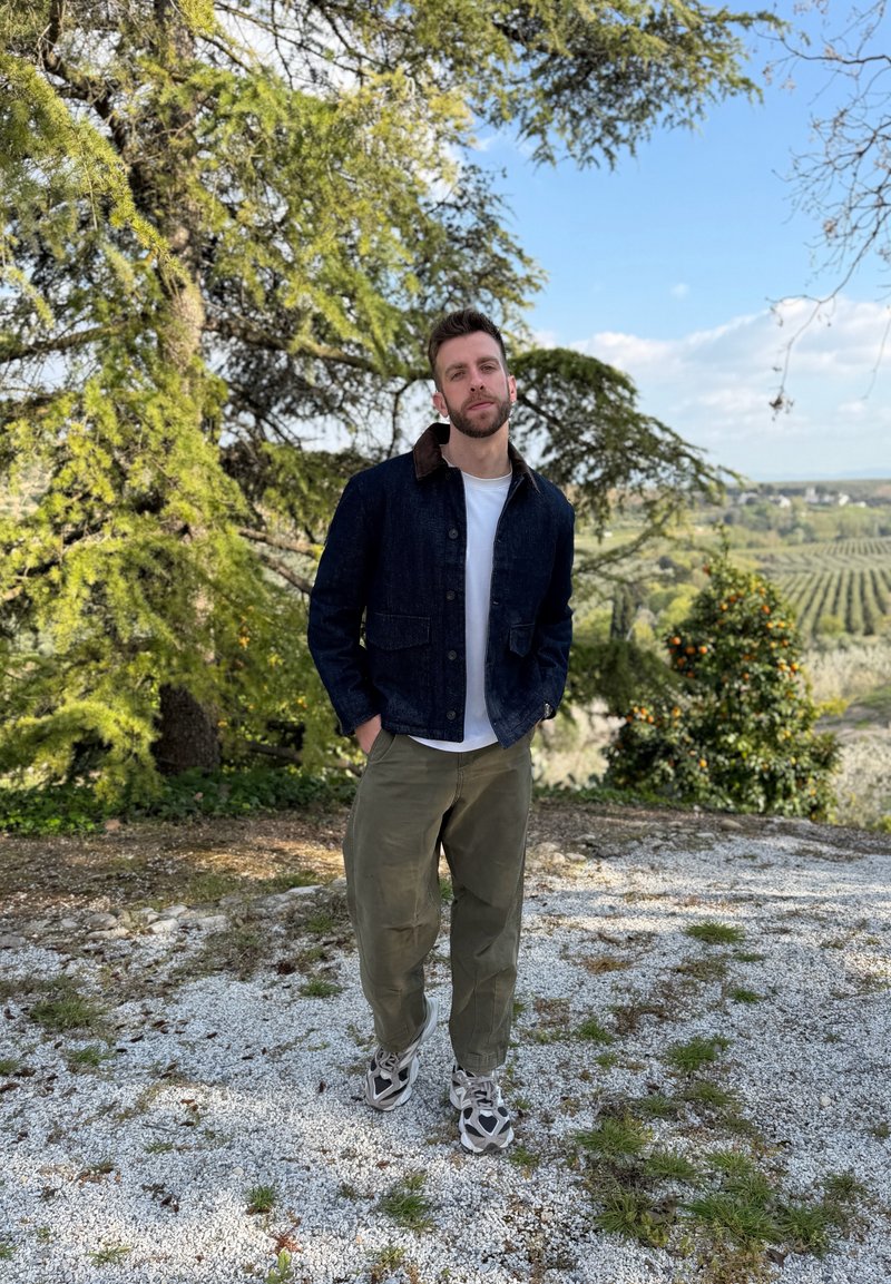 Man wearing denim jacket, white shirt, and olive pants stands on gravel path near trees with a vineyard in the background under a blue sky.