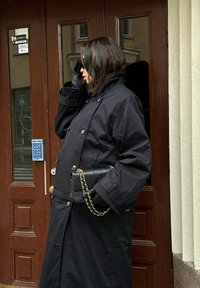 Black knee-length coat featuring a high collar and large buttons. Accompanied by leather gloves and a quilted black handbag with a gold chain strap.