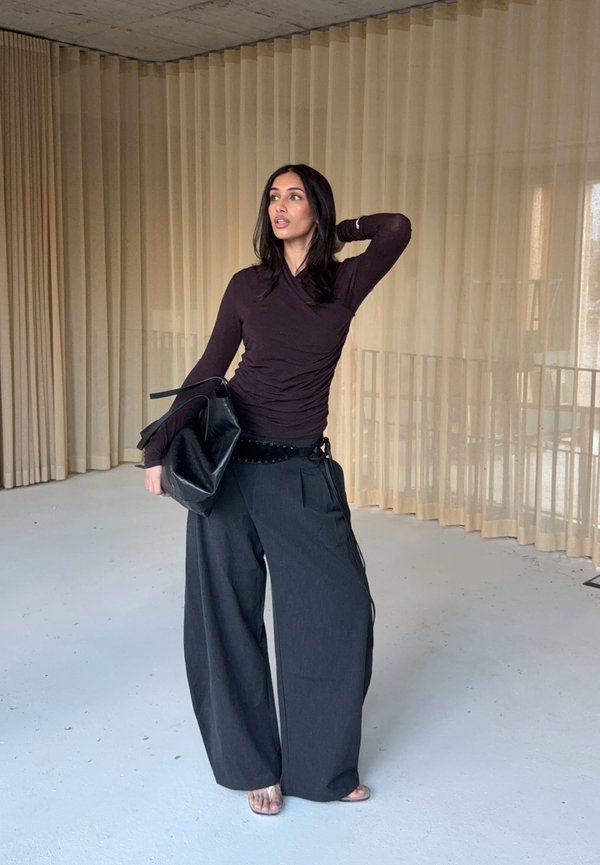 Um modelo veste uma blusa de mangas compridas justas castanho-escuro e calças largas pretas, segurando uma bolsa tote preta. Texturas simples e suaves.