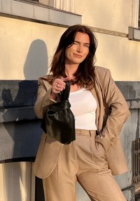 Costume beige sur mesure avec larges revers, haut blanc ajusté, tenant un sac à main en cuir noir à la texture douce, devant un mur texturé.