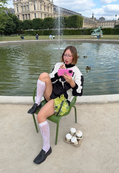 Persona sentada en una silla verde al lado de un estanque, vestida con una chaqueta blanca y negra, medias de encaje blancas y zapatos negros, con un teléfono rosa y una bolsa verde.